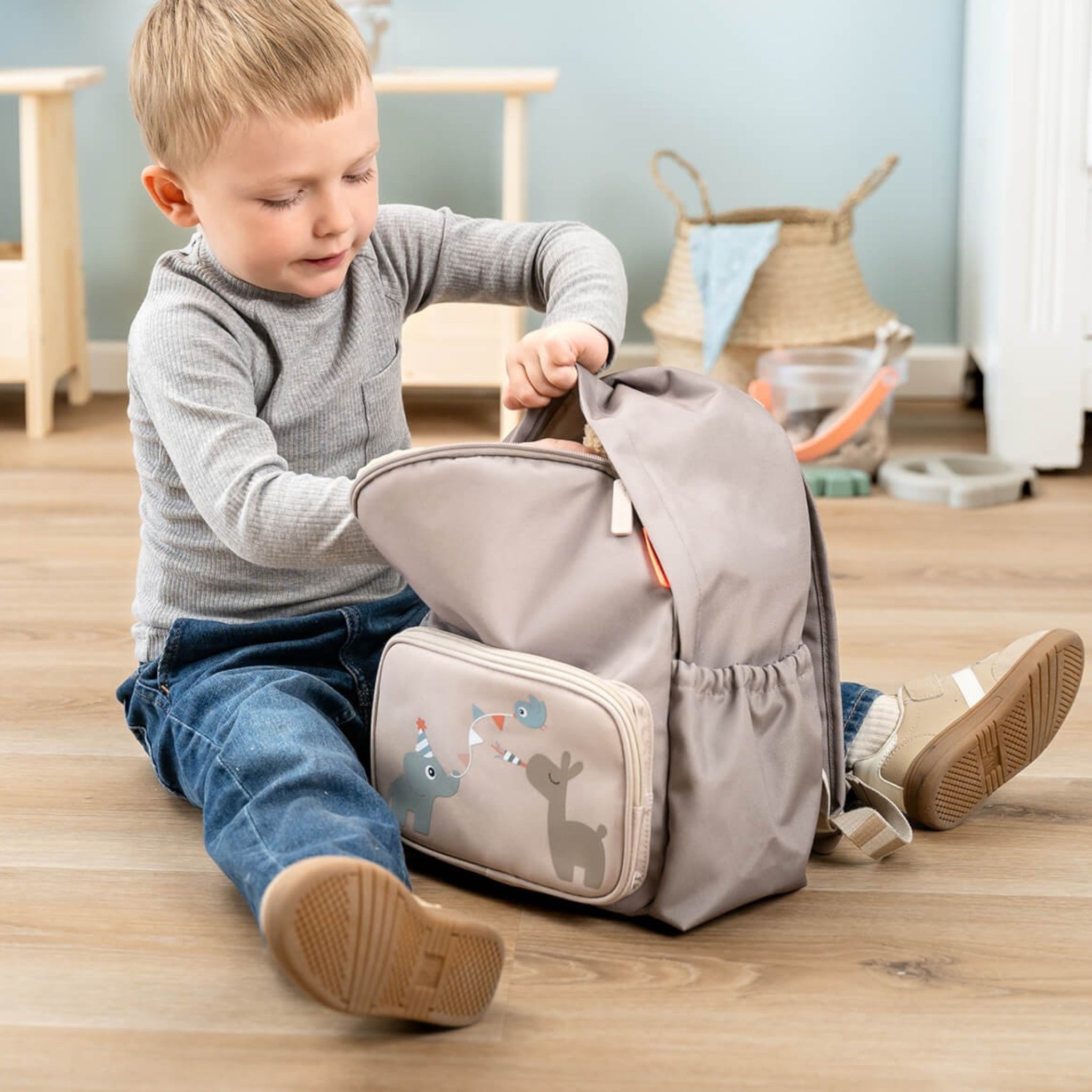 Young boy sitting on a wooden floor opening a sand-colored Lil Bunnies - The Collective kids backpack with animal art