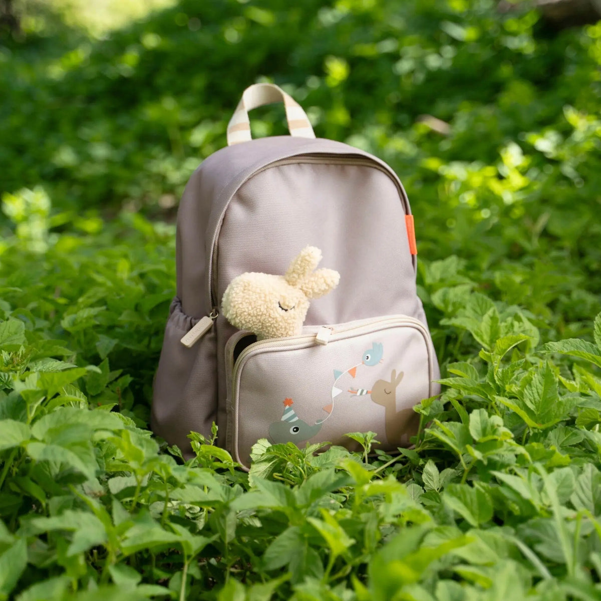 Lil Bunnies - The Collective sand-colored kids backpack with a plush bunny toy in the front pocket, set in greenery.