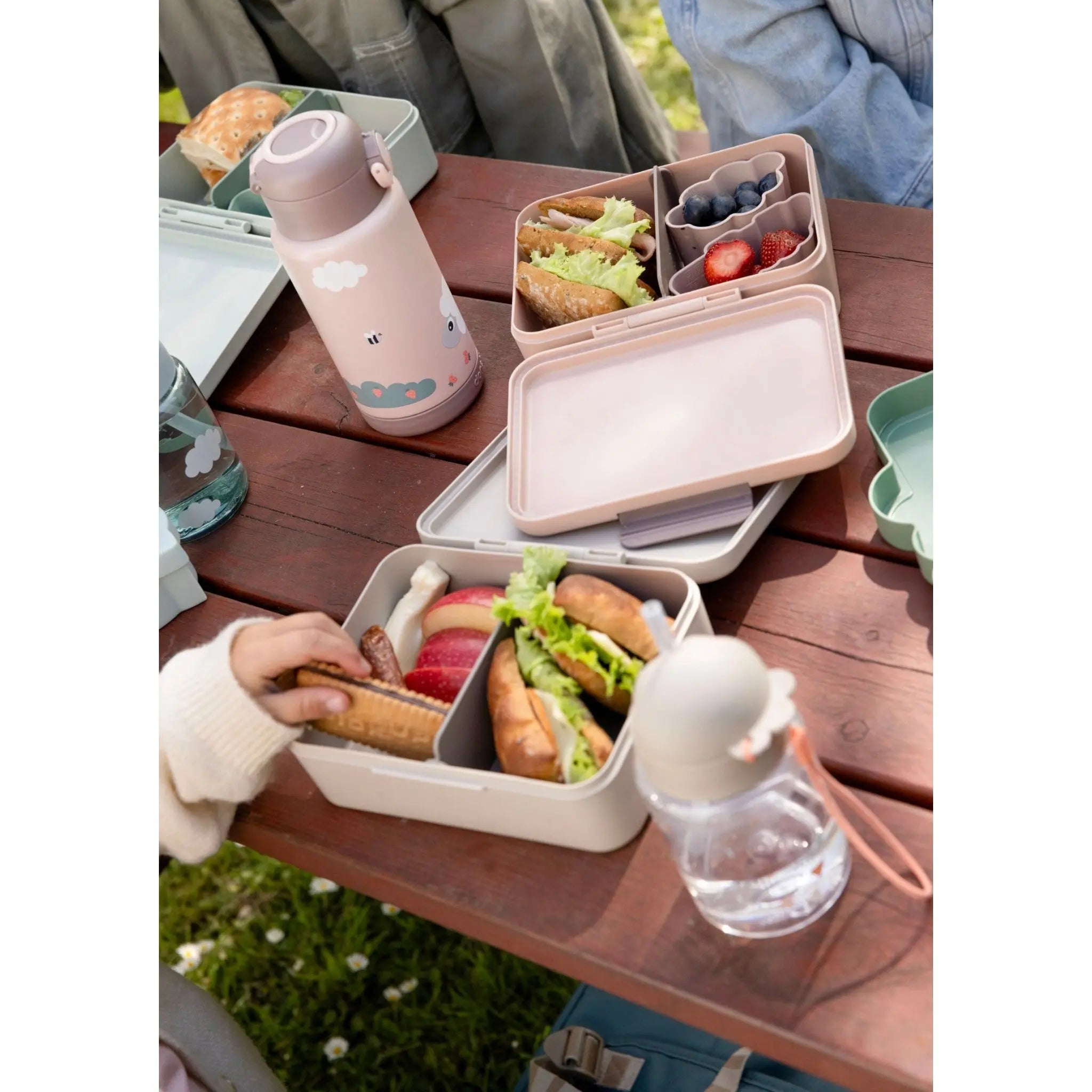Lil Bunnies - The Collective sand-colored lunch box with divider, filled with sandwiches and fruit on a picnic table.