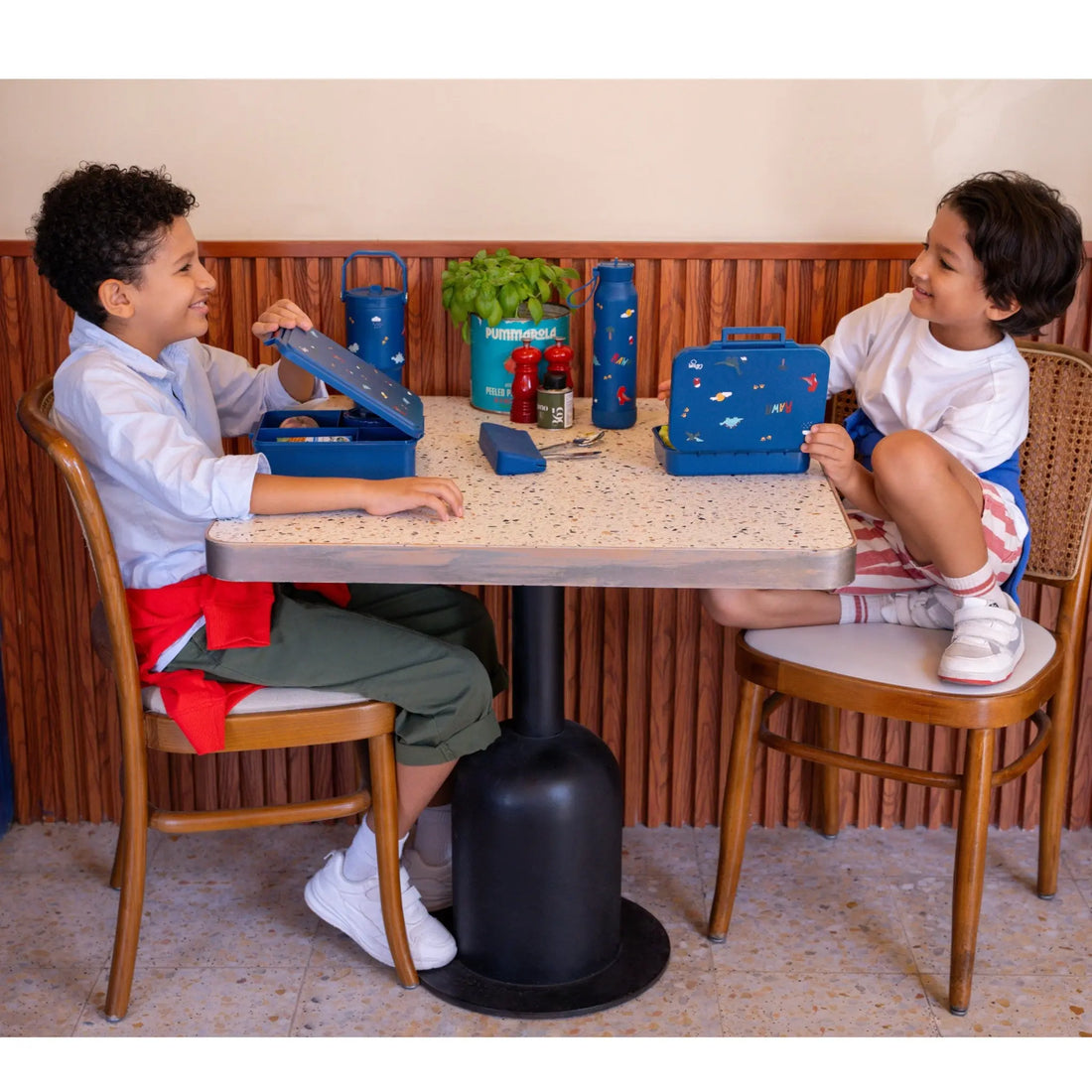 Two children sitting at a table with Lil Bunnies - The Collective blue dinosaur print lunchboxes and food jars.