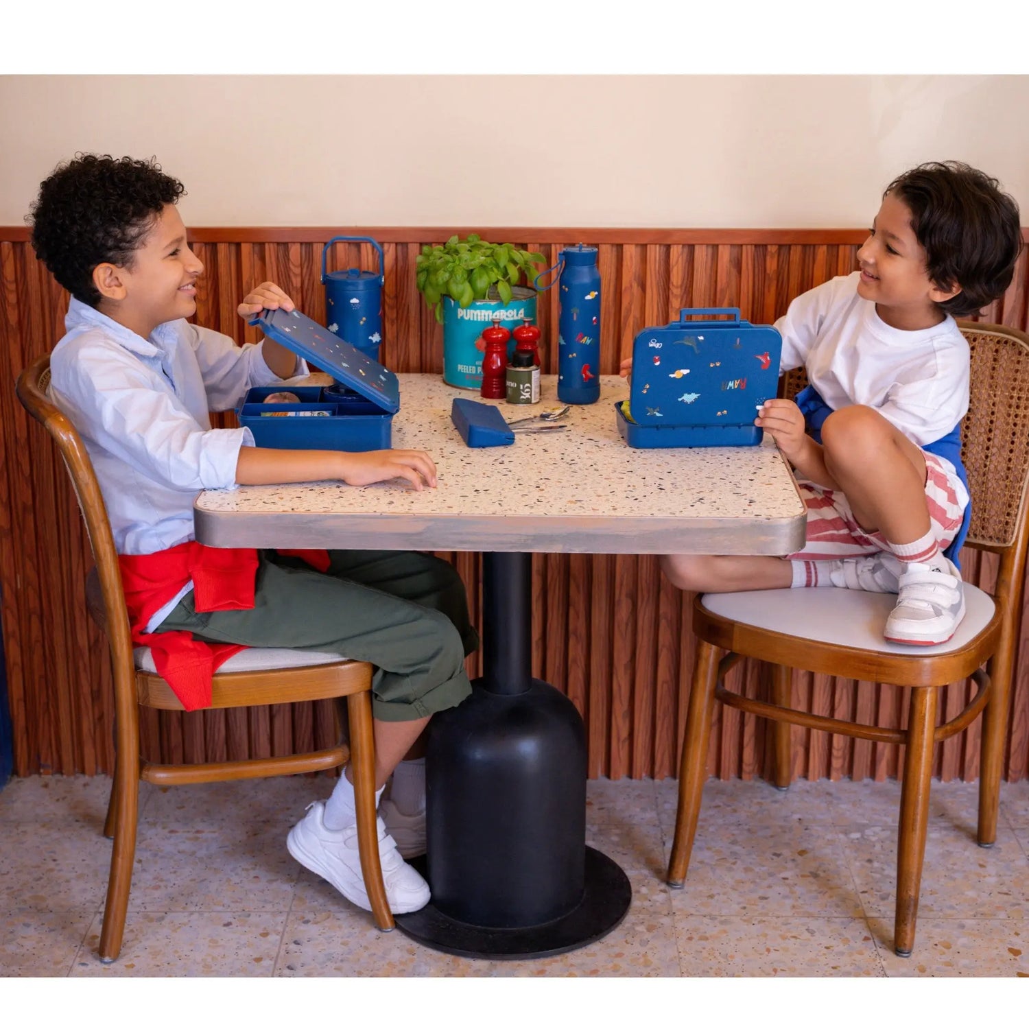 Two children sitting at a table with Lil Bunnies - The Collective blue dinosaur print lunchboxes and food jars.