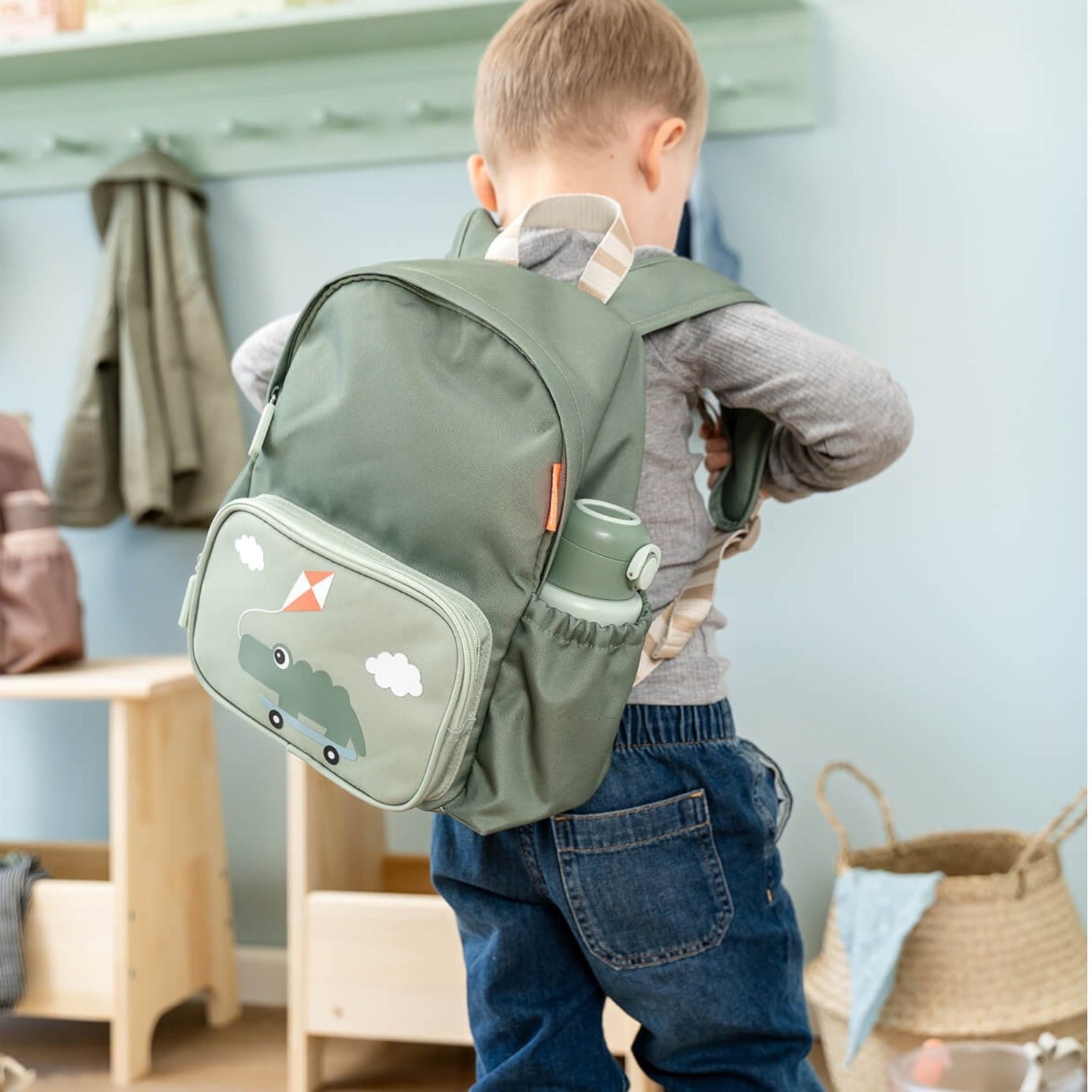 A young boy wearing a sage green Lil Bunnies - The Collective kids backpack with a cute dinosaur and kite print.