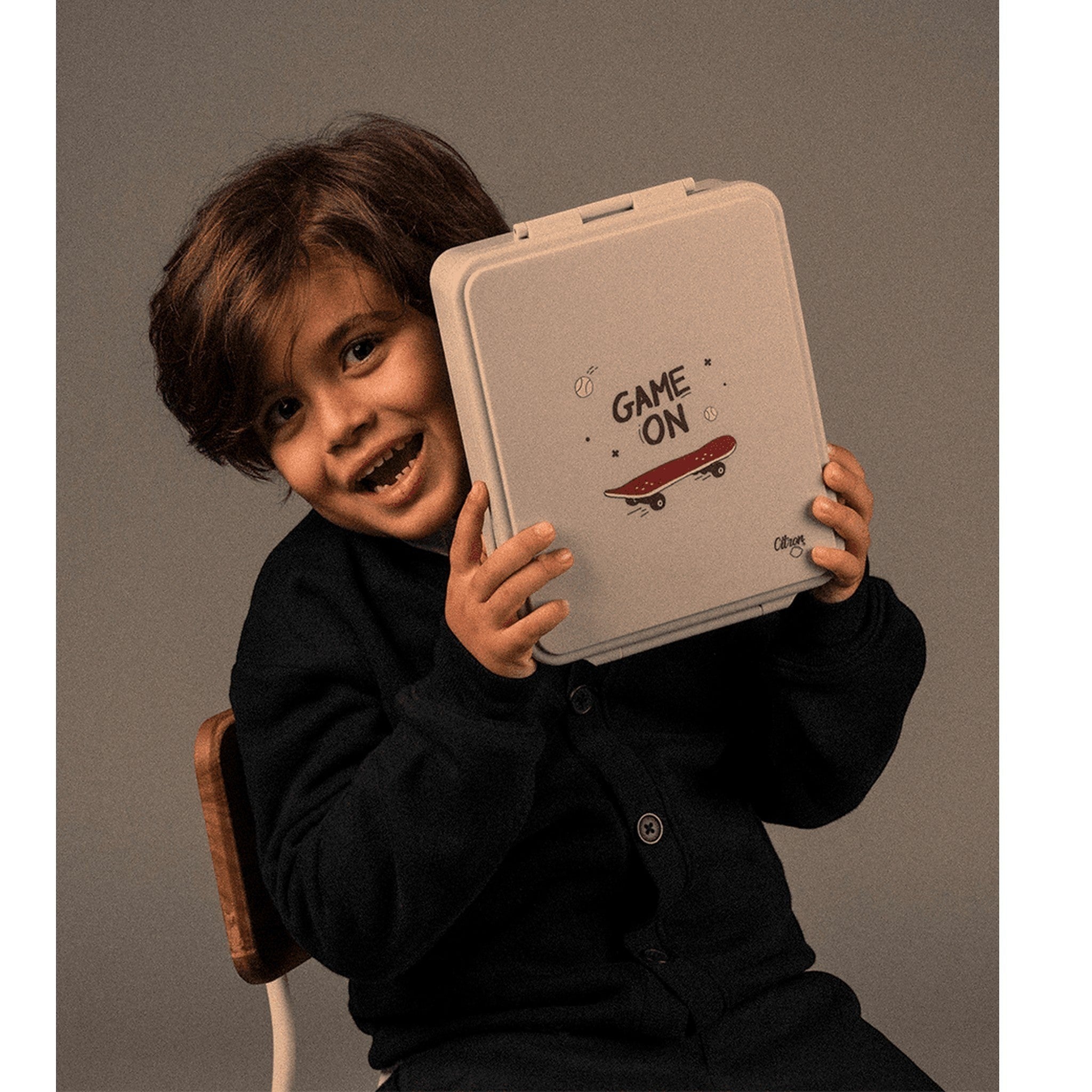 A smiling boy holding a Lil Bunnies - The Collective grey lunch box with a Game On skateboard graphic