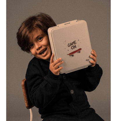 A smiling boy holding a Lil Bunnies - The Collective grey lunch box with a Game On skateboard graphic
