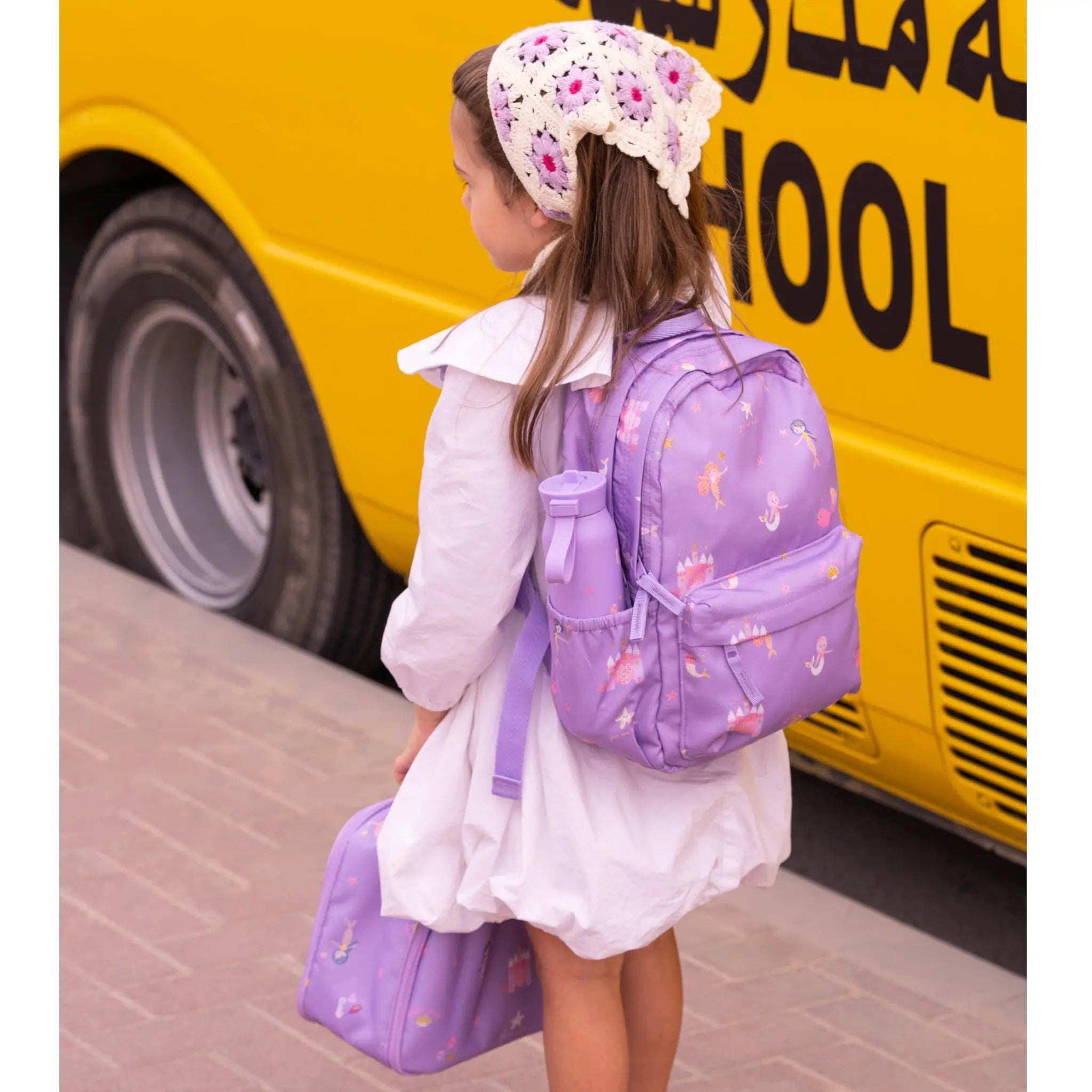 A young girl wearing a Lil Bunnies - The Collective purple mermaid print backpack and matching lunch bag by a bus