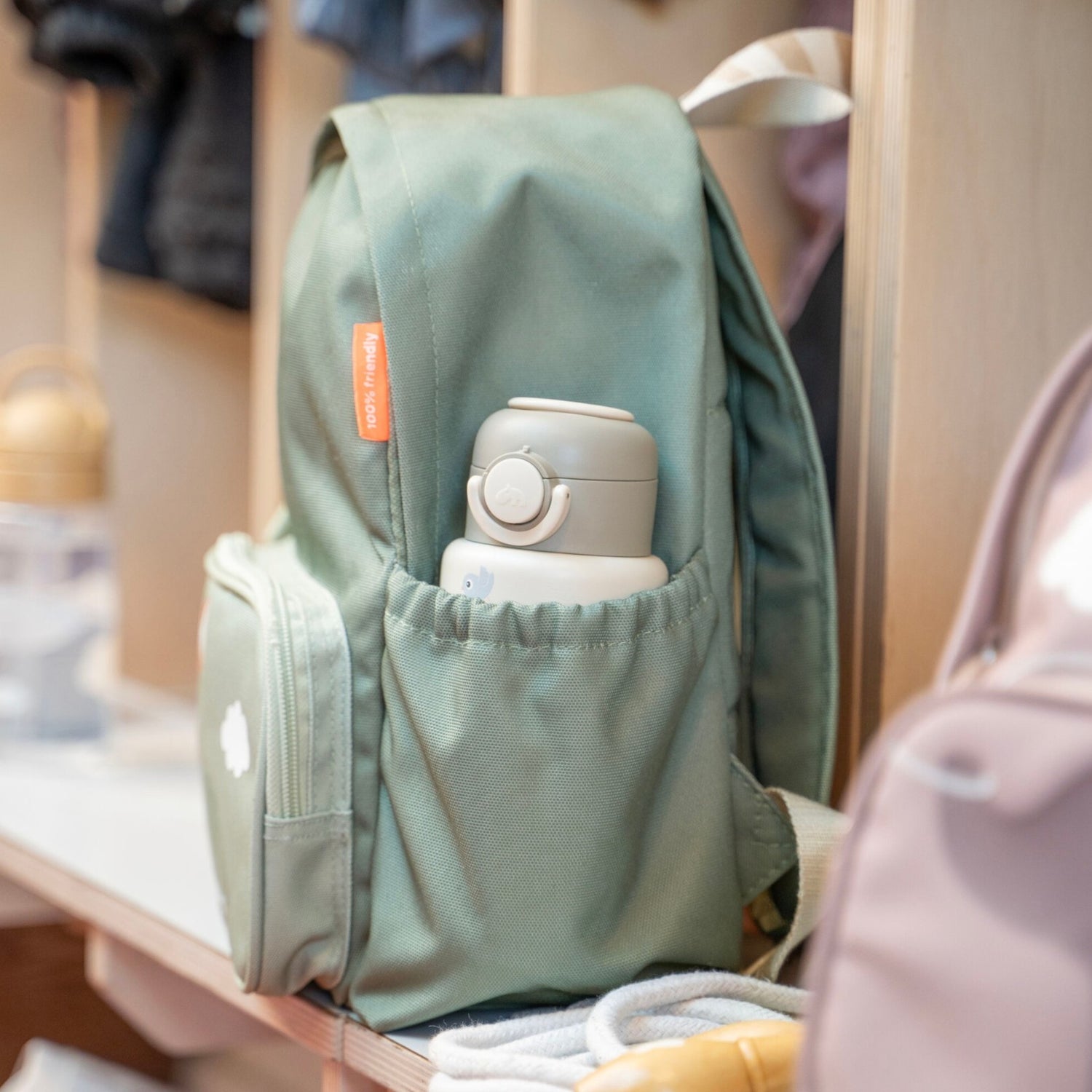 Lil Bunnies - The Collective sand-colored insulated bottle tucked into the side pocket of a sage green backpack
