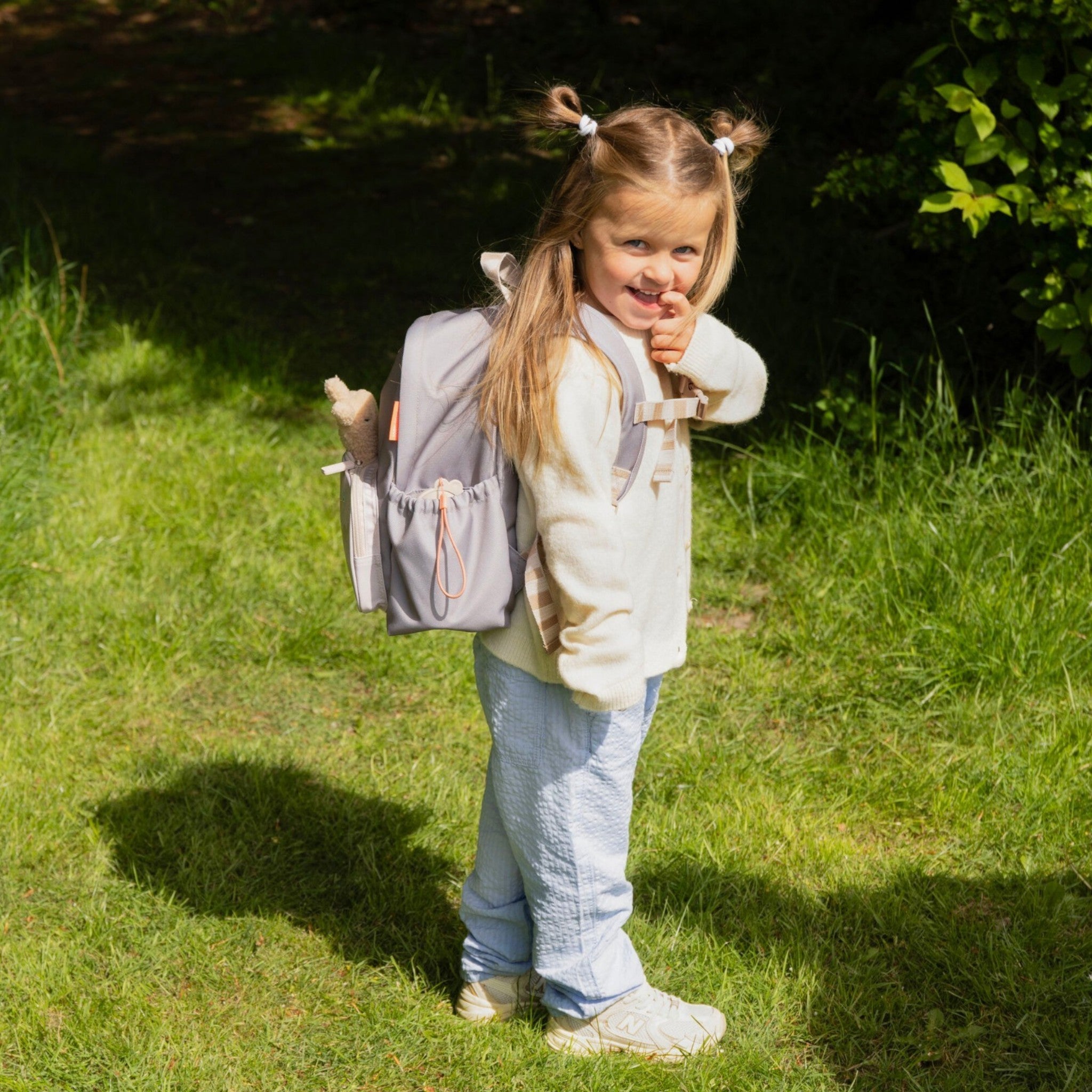 A young girl wearing a sand-colored Lil Bunnies - The Collective backpack while standing in a sunny grassy park.