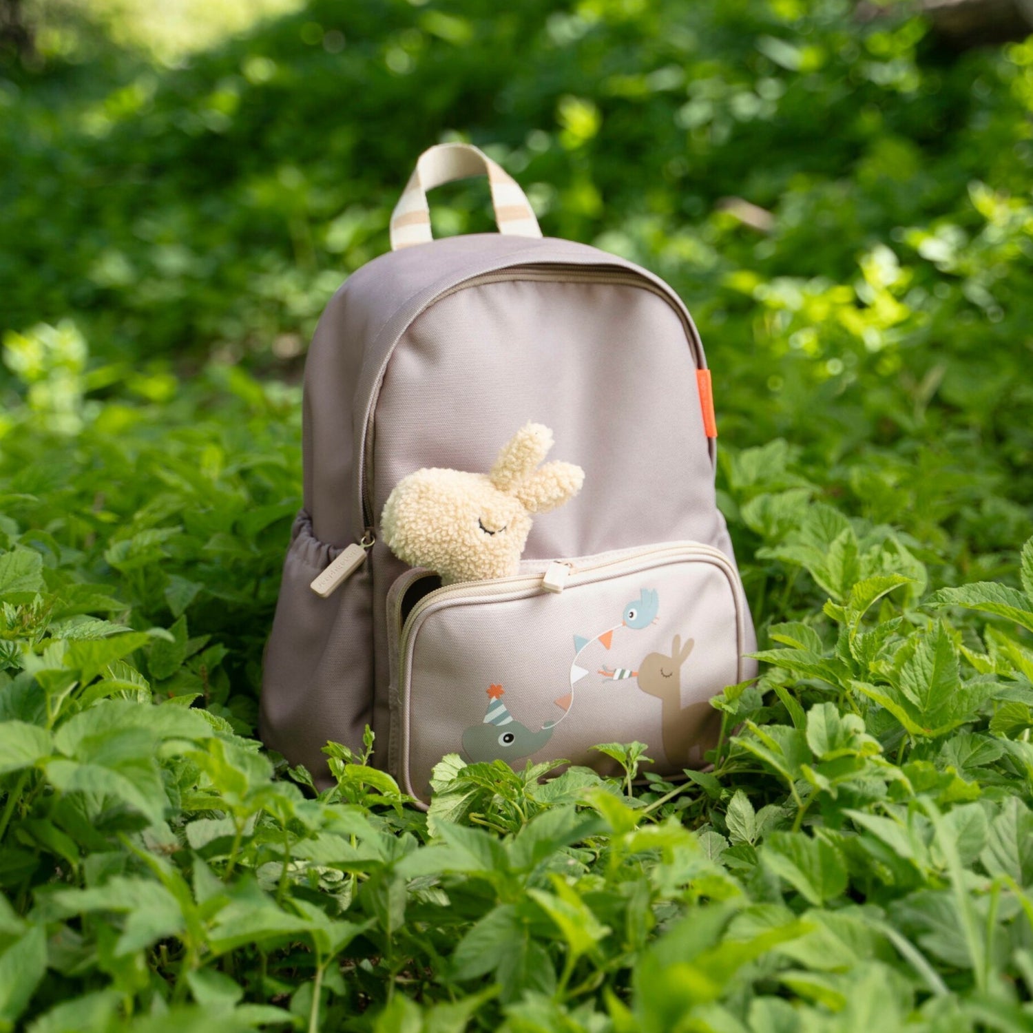 Lil Bunnies - The Collective sand-colored kids backpack with a plush bunny toy in the front pocket, set in greenery.
