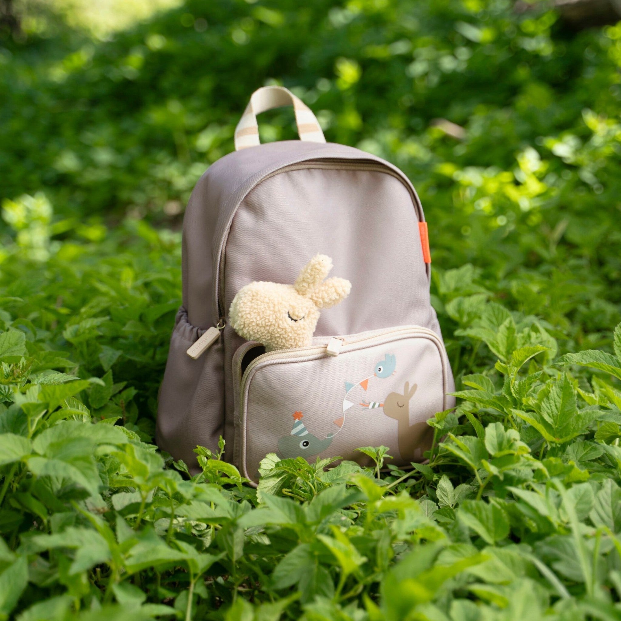 Lil Bunnies - The Collective sand-colored kids backpack with a plush bunny toy in the front pocket, set in greenery.