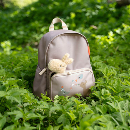 Lil Bunnies - The Collective sand-colored kids backpack with a plush bunny toy in the front pocket, set in greenery.