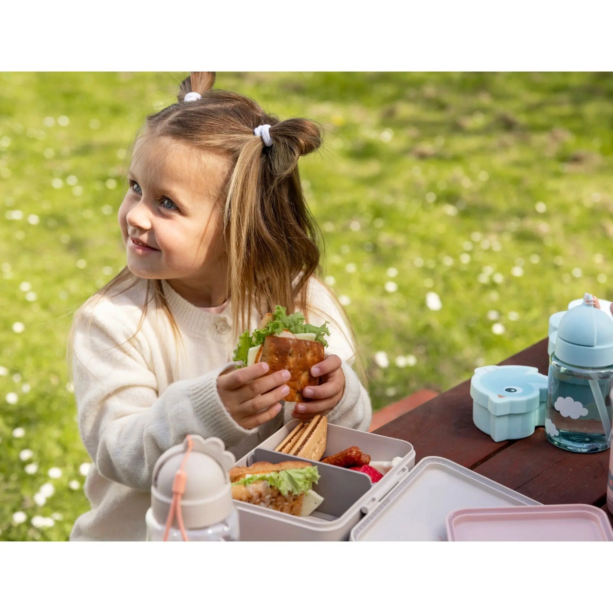 Lil Bunnies - The Collective sand-coloured lunch box with divider, filled with a sandwich, on a picnic table.