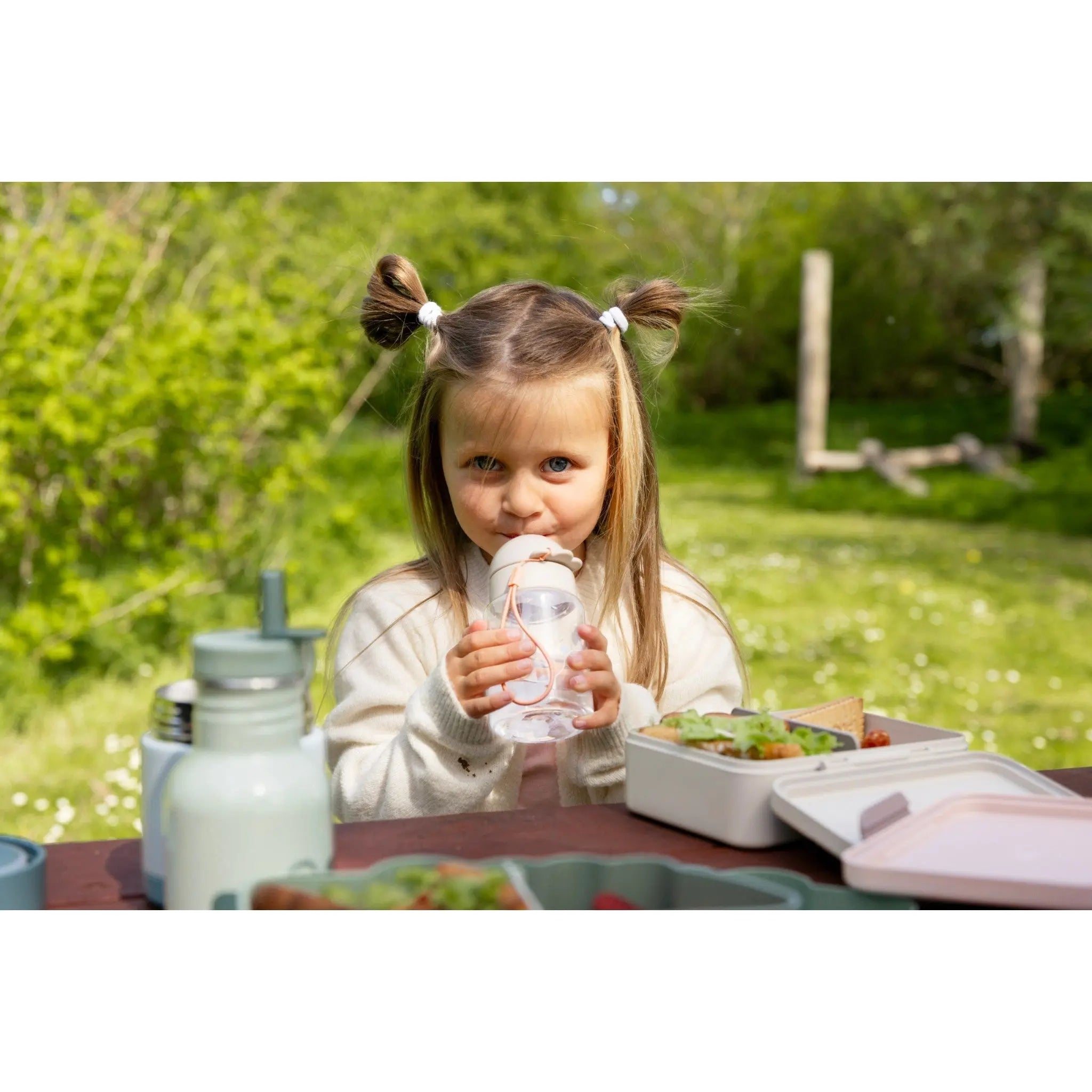 Lil Bunnies - The Collective sand-coloured lunch box with divider on a picnic table with a young girl drinking water