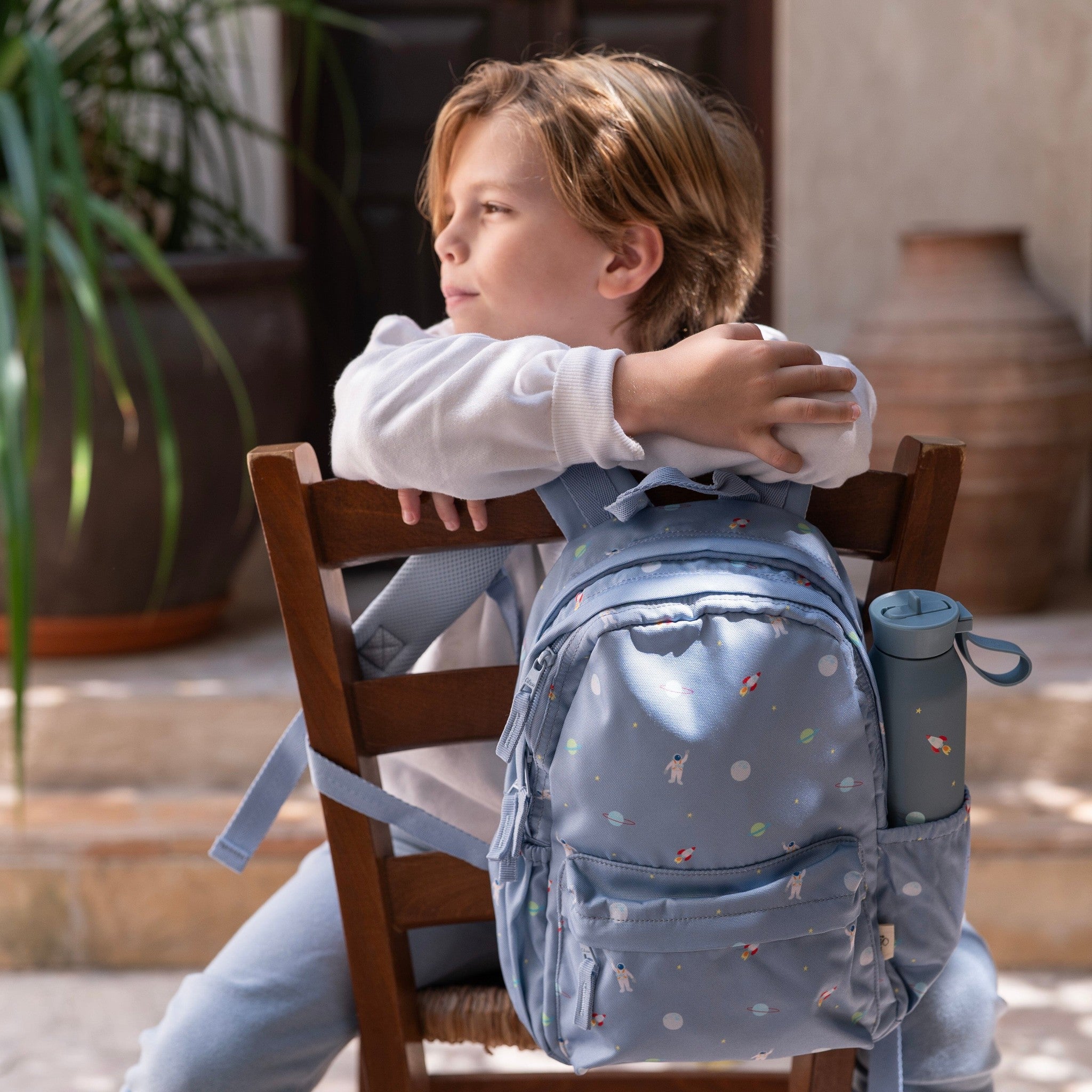 Lil Bunnies - The Collective medium blue backpack with a spaceship print, worn by a child sitting on a wooden chair.